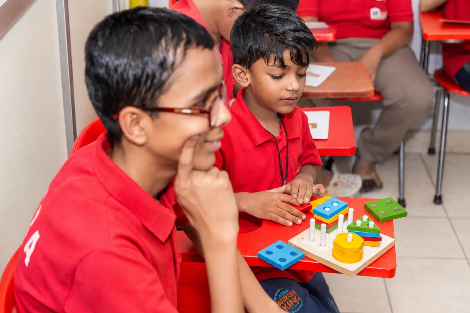 Two children in red HCRA uniforms playing with a colorful wooden shape-sorting puzzle in a special education classroom